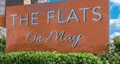 A sign that says "The Flats One Way" in front of a hedge.